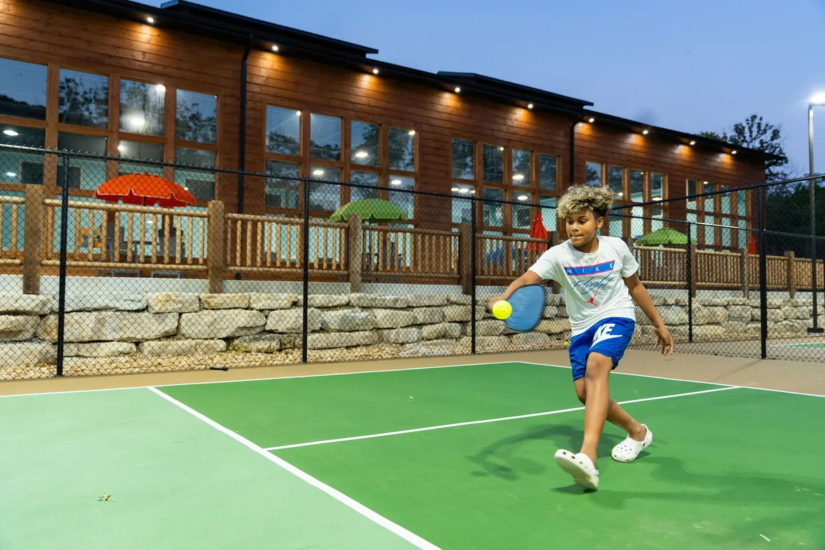 A close-up of a child playing pickleball on a dedicated court, with colorful umbrellas and a modern wooden building in the background. The action captures a playful, energetic moment, surrounded by an inviting, well-maintained recreational space.