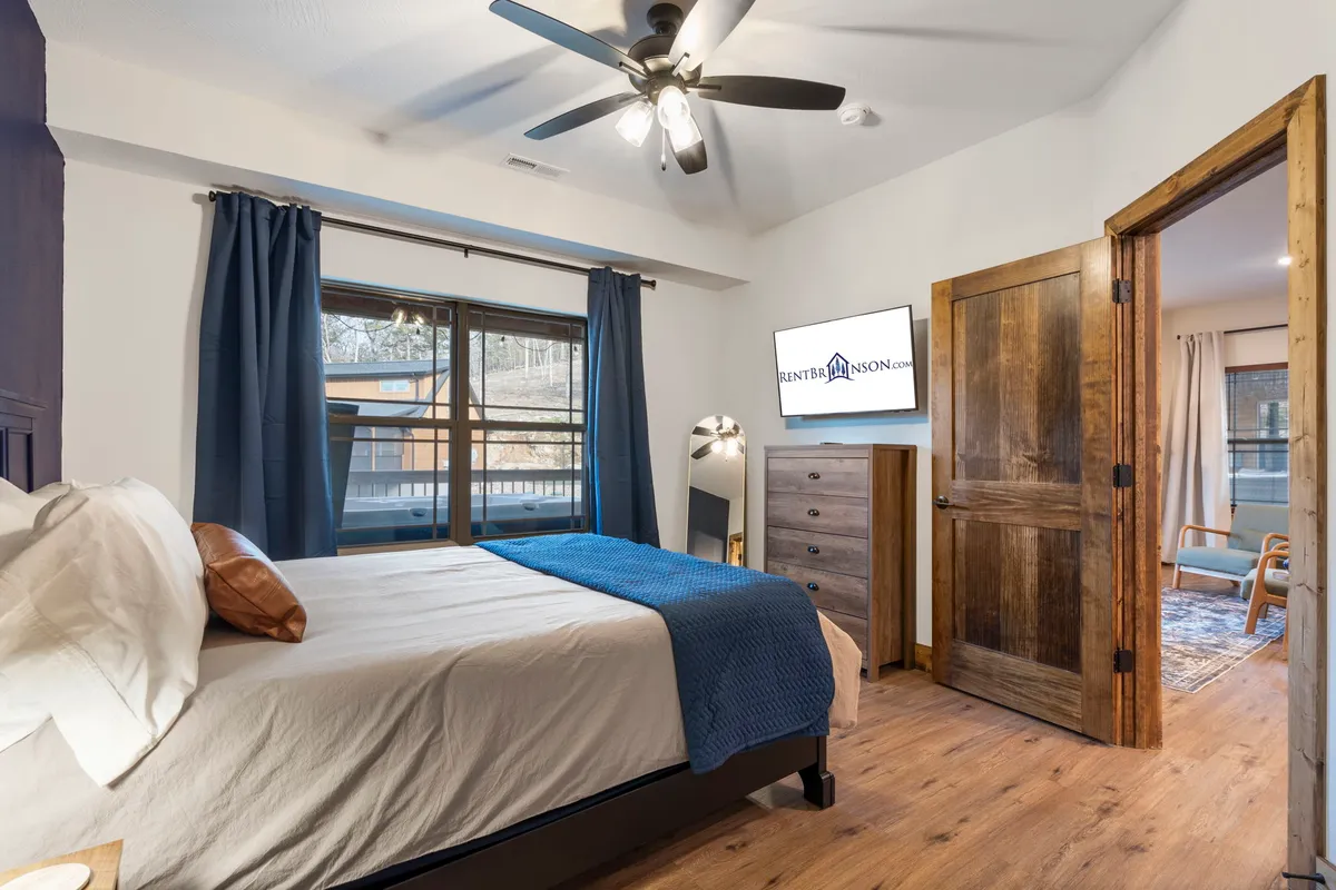 A cozy bedroom featuring a bed with a blue throw, a window with curtains letting in soft natural light, a wooden dresser, and a television across from the bed. The room feels inviting and perfect for relaxing or catching up on your favorite shows.