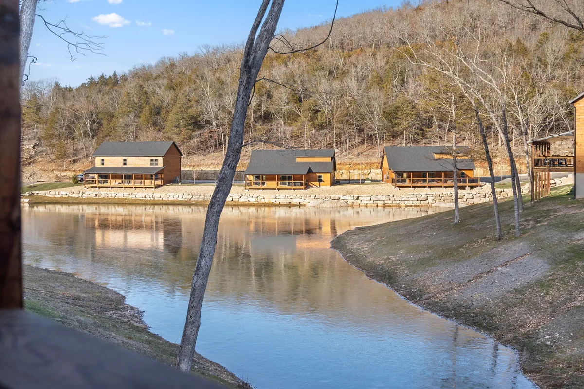 A tranquil lakeside scene with cabins reflected on calm water, framed by trees lining the shore. The ambiance is serene and inviting, perfect for fishing, kayaking, or simply soaking in the beauty of the stocked pond.