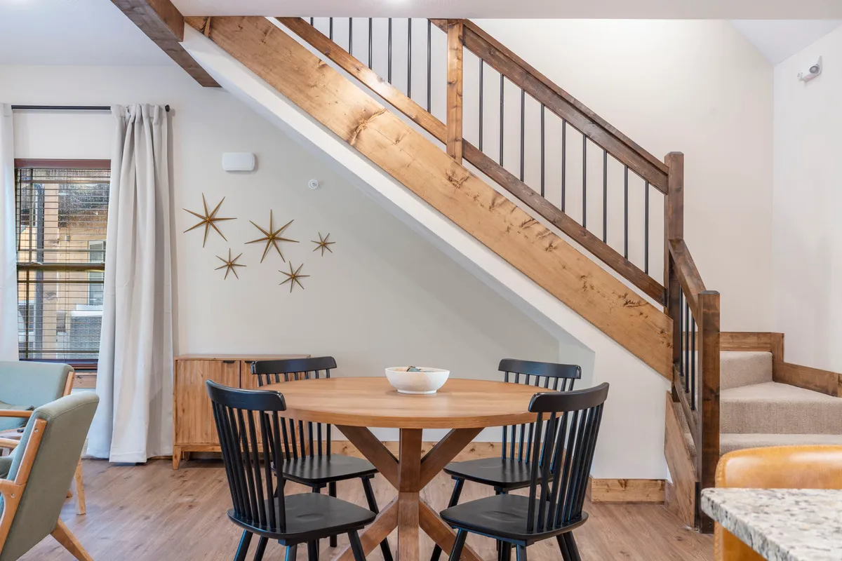 A round wooden dining table surrounded by black dining chairs sits beneath decorative wall stars, adjacent to a wooden staircase. The setting radiates a cozy, intimate vibe, ideal for shared meals and game nights.