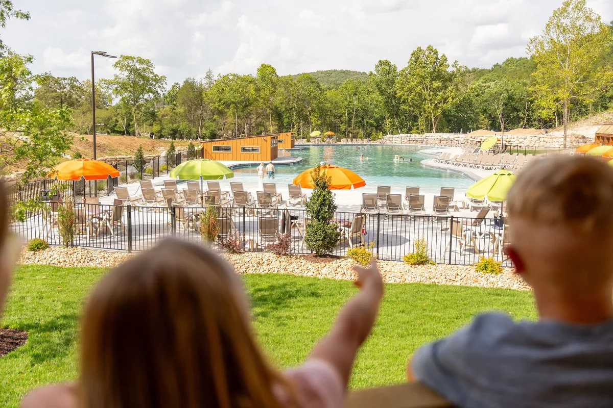 An outdoor swimming pool bustling with activity, surrounded by colorful umbrellas and children happily pointing and playing. The setting is bathed in sunlight, with families enjoying poolside gatherings and laughter echoing across the water.