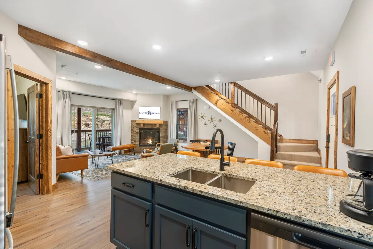 A bright, open-concept living space that seamlessly combines a granite kitchen island, comfortable seating area, fireplace, and a staircase leading to the upper level. The large windows flood the area with natural light, enhancing the welcoming ambiance and offering views outside.
