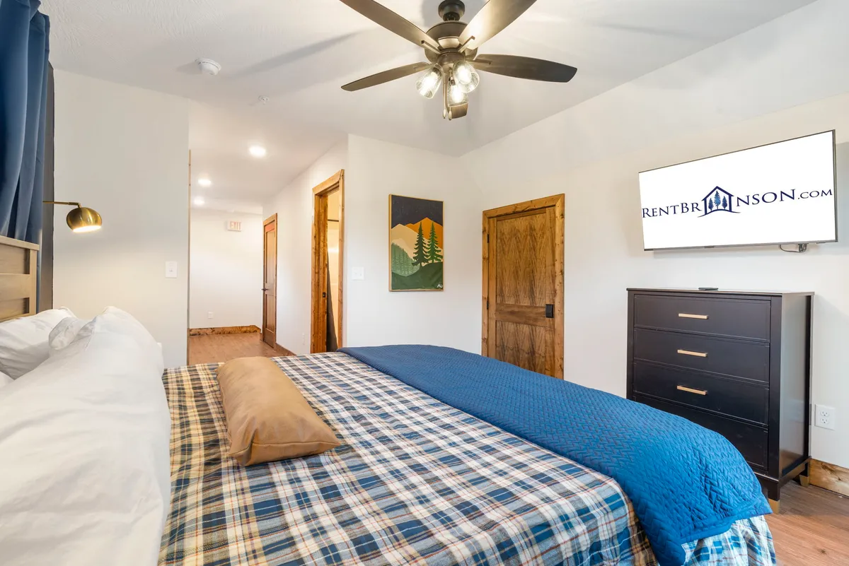 A comfortable bedroom with plaid bedding, decorative pillows, a TV mounted on the wall, artwork, and a dresser. The overall ambiance is homely and restful, ideal for relaxation and quiet moments.