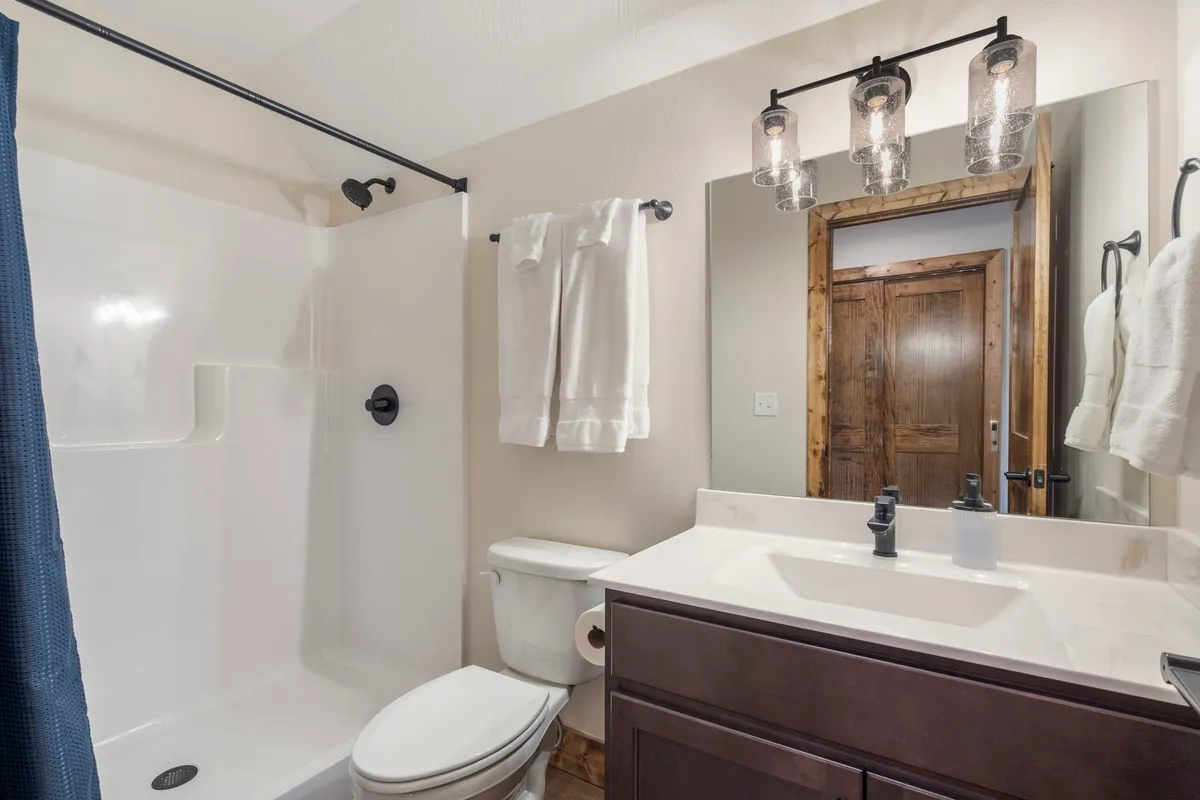 A modern ensuite bathroom with a spacious vanity and sink, a walk-in shower area, toilet, mirror, and stylish light fixtures. The space feels clean, serene, and ideal for restorative routines.