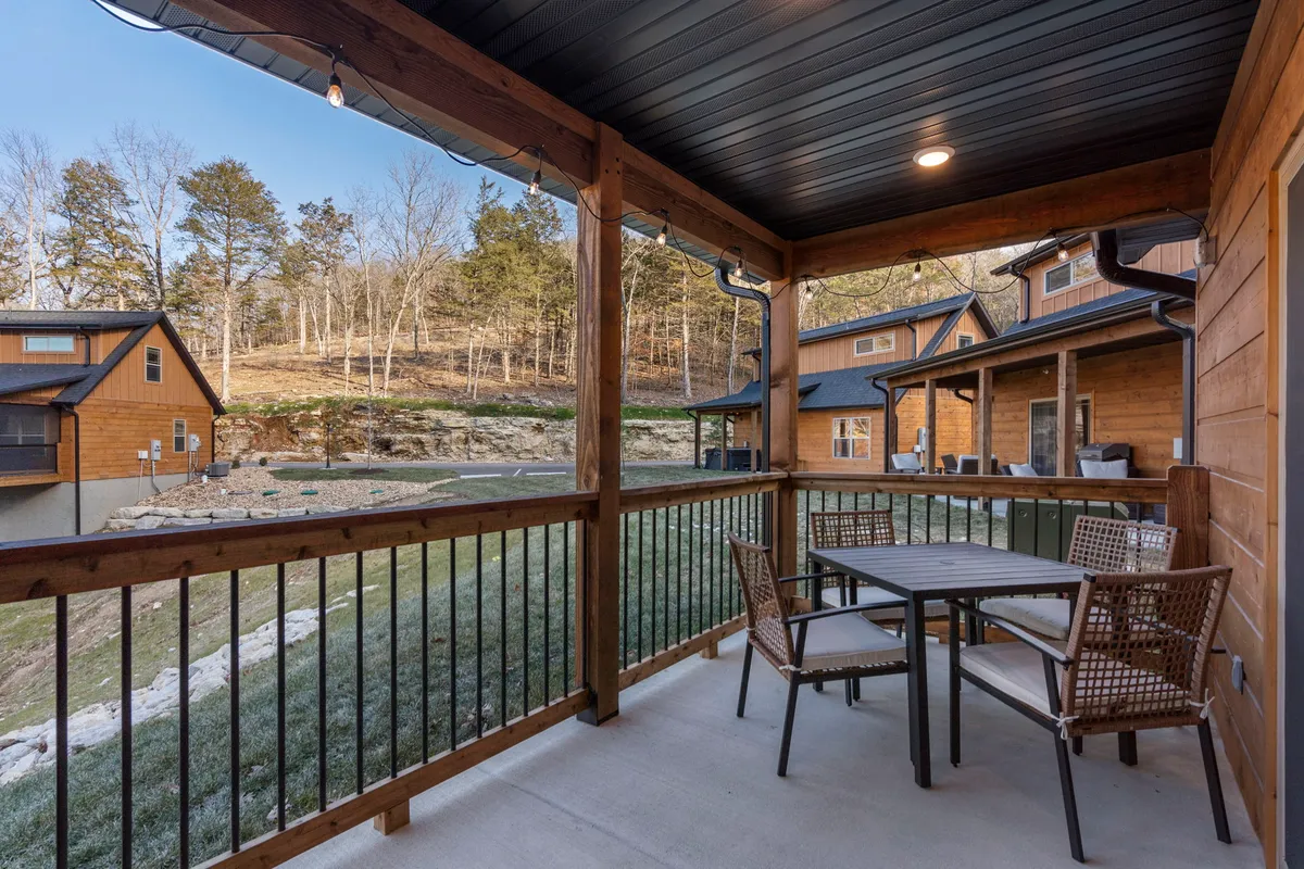 A peaceful exterior patio scene featuring a patio table surrounded by lush trees and a wooden railing. The setting invites morning coffee, outdoor dining, or quiet conversation, with nature closely embracing the seating area.