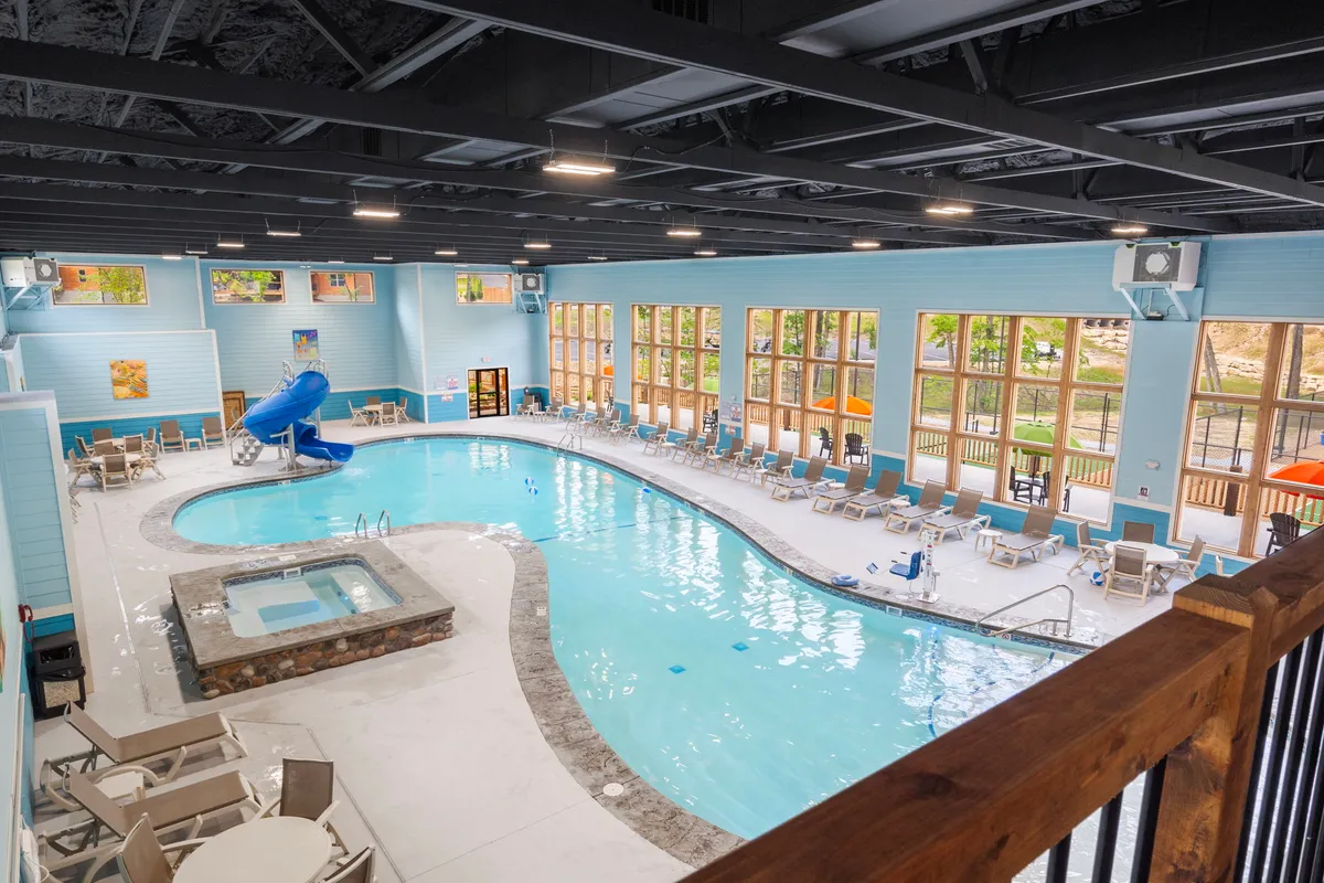 A vibrant indoor swimming pool area filled with natural light, featuring a water slide, spa area, and plenty of poolside lounge chairs. Children are seen playing on the water slide while parents relax in the spacious setting, which is accentuated by large windows offering views of the outdoors.