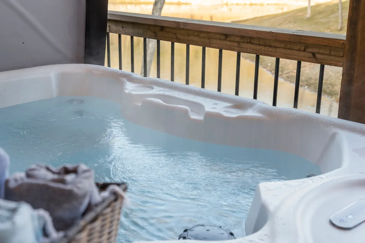 A tranquil indoor hot tub area with a basket of towels placed nearby. Through the large window, soft evening light filters in, and the space is styled to evoke a serene, spa-like experience surrounded by nature.