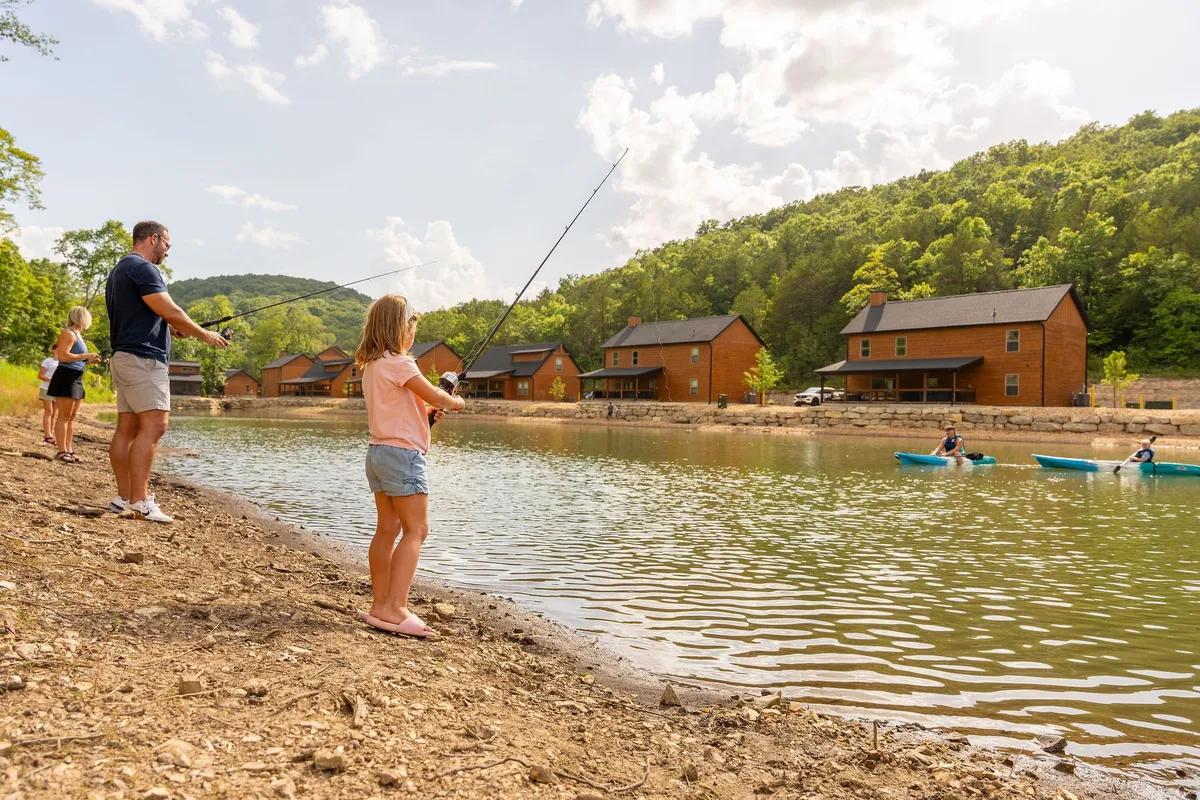 A lively exterior scene with families fishing, kayakers on the water, and wooden cabins in the background. The lush greenery and calm water set a playful and serene mood, emphasizing outdoor adventure and family bonding.