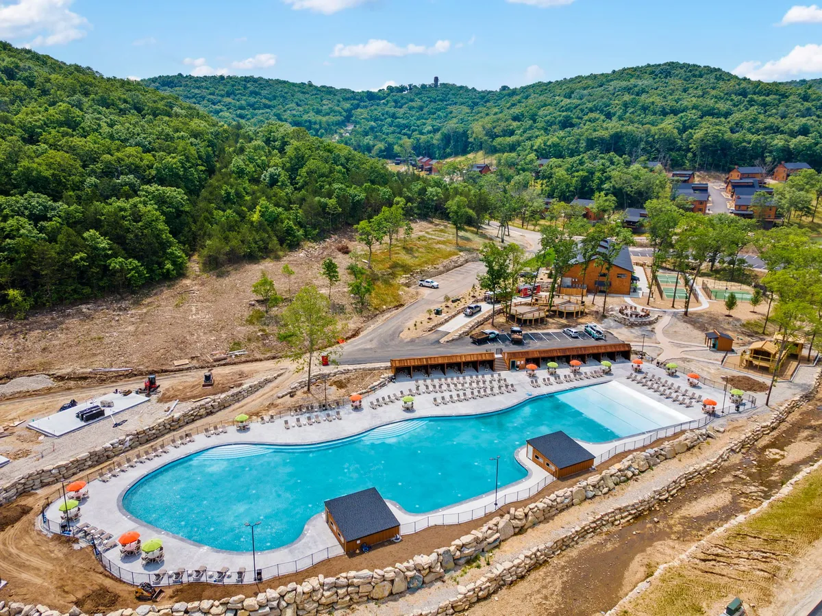 An inviting view of a large outdoor swimming pool surrounded by lounge chairs, cabana-like structures, and a forested backdrop. Pathways lead to the pool, and the whole area is bathed in warm, natural light, suggesting relaxation and family fun.