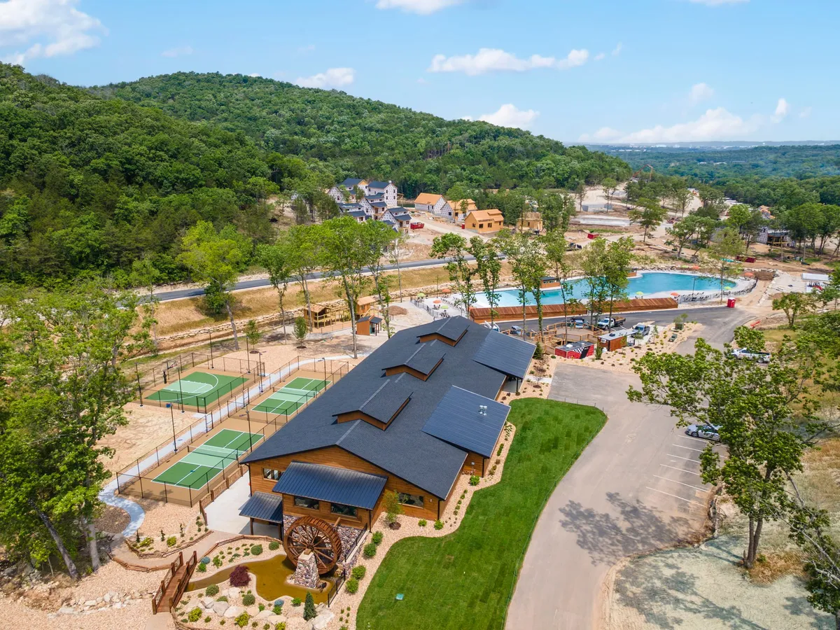 A wide exterior shot of Tall Timbers Camp's main building, surrounded by vibrant tennis courts, a sparkling swimming pool, a distinctive wheel feature, and scenic walking trails. The scene is lively, with guests enjoying outdoor amenities and the landscape dotted with greenery.