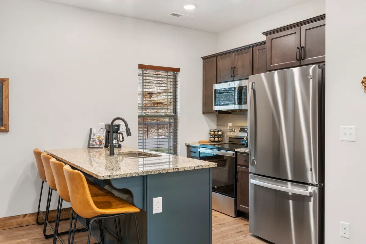 A kitchen scene featuring a granite countertop, bar stools for casual conversation, stainless steel appliances, and a window streaming in natural light. The layout is functional yet welcoming, ideal for culinary adventures or casual snacks.