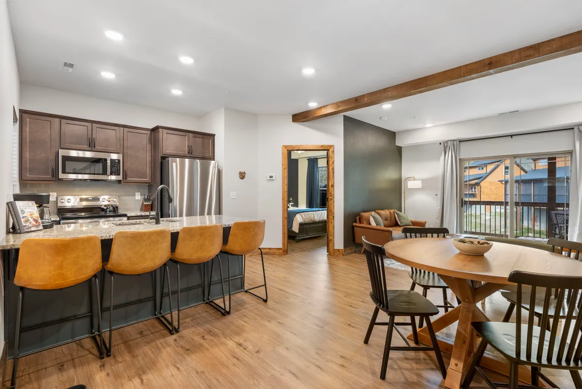 The image features a warm, inviting cabin interior where a spacious kitchen island with bar stools seamlessly flows into a cozy dining area. An open doorway leads to a tranquil bedroom, while the scene exudes a sense of togetherness, perfect for sharing meals and stories in a modern yet rustic setting.